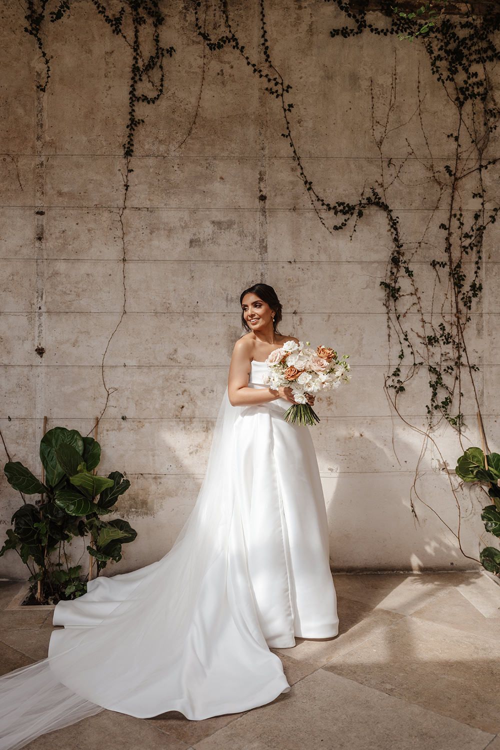 Elegant bride in flowing white gown poses against a vine-covered wall while holding a lush bouquet of flowers.