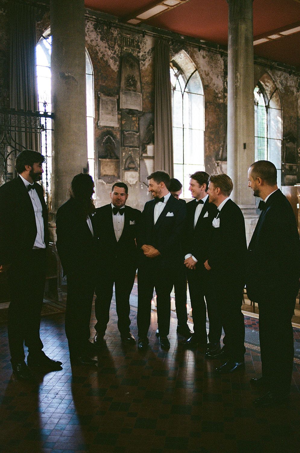 A line of groomsmen in black tuxedos stand together in a dimly lit hallway with arched windows.