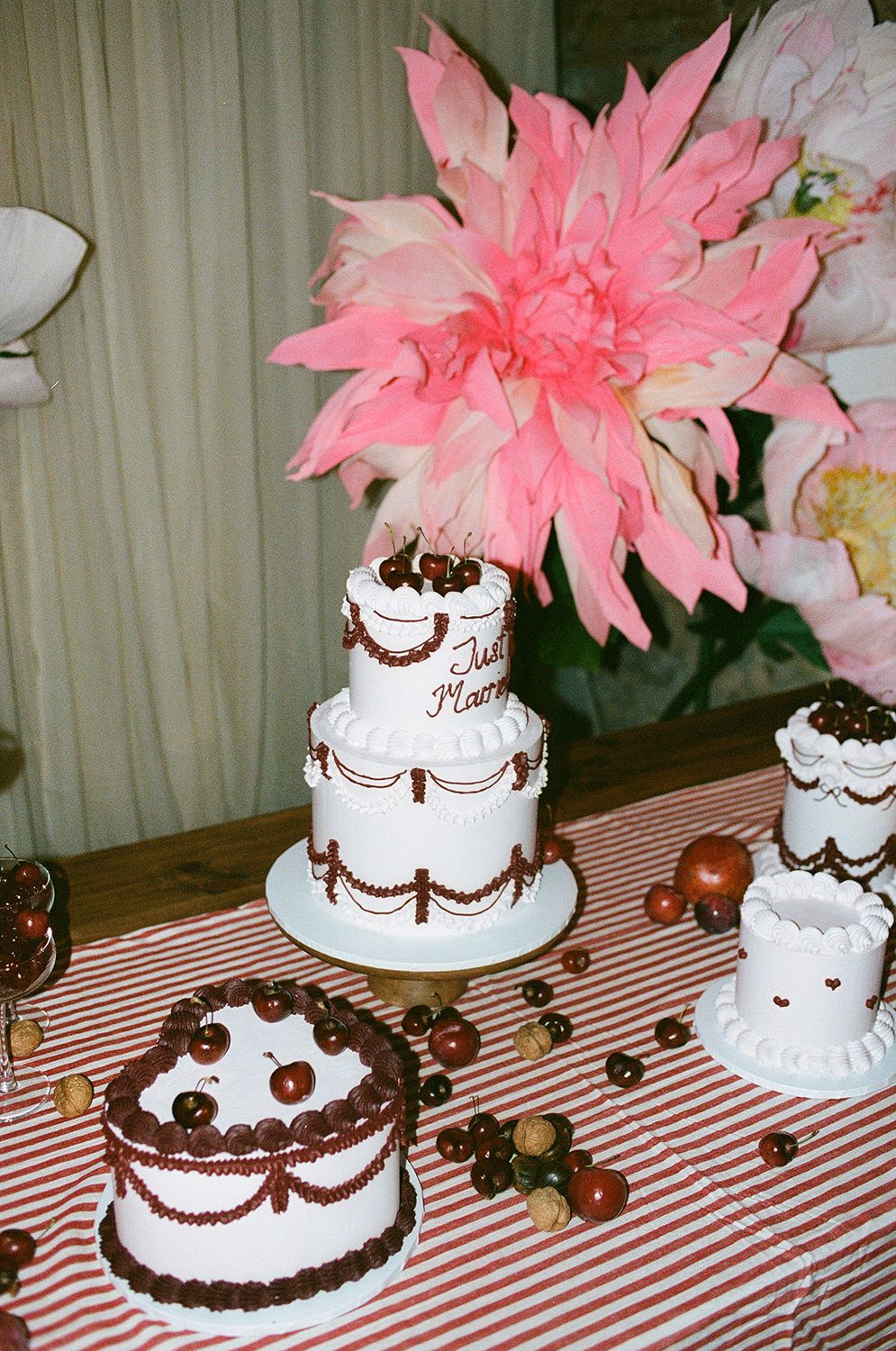Pink floral arrangement with white tiered cakes decorated with brown piping and truffles on a red striped tablecloth.