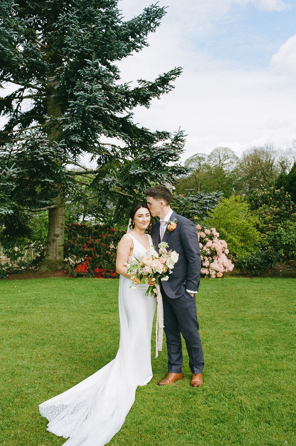 Bride and groom stood smiling in the gardens at The Holford Estate, the bride is holding her flowers.