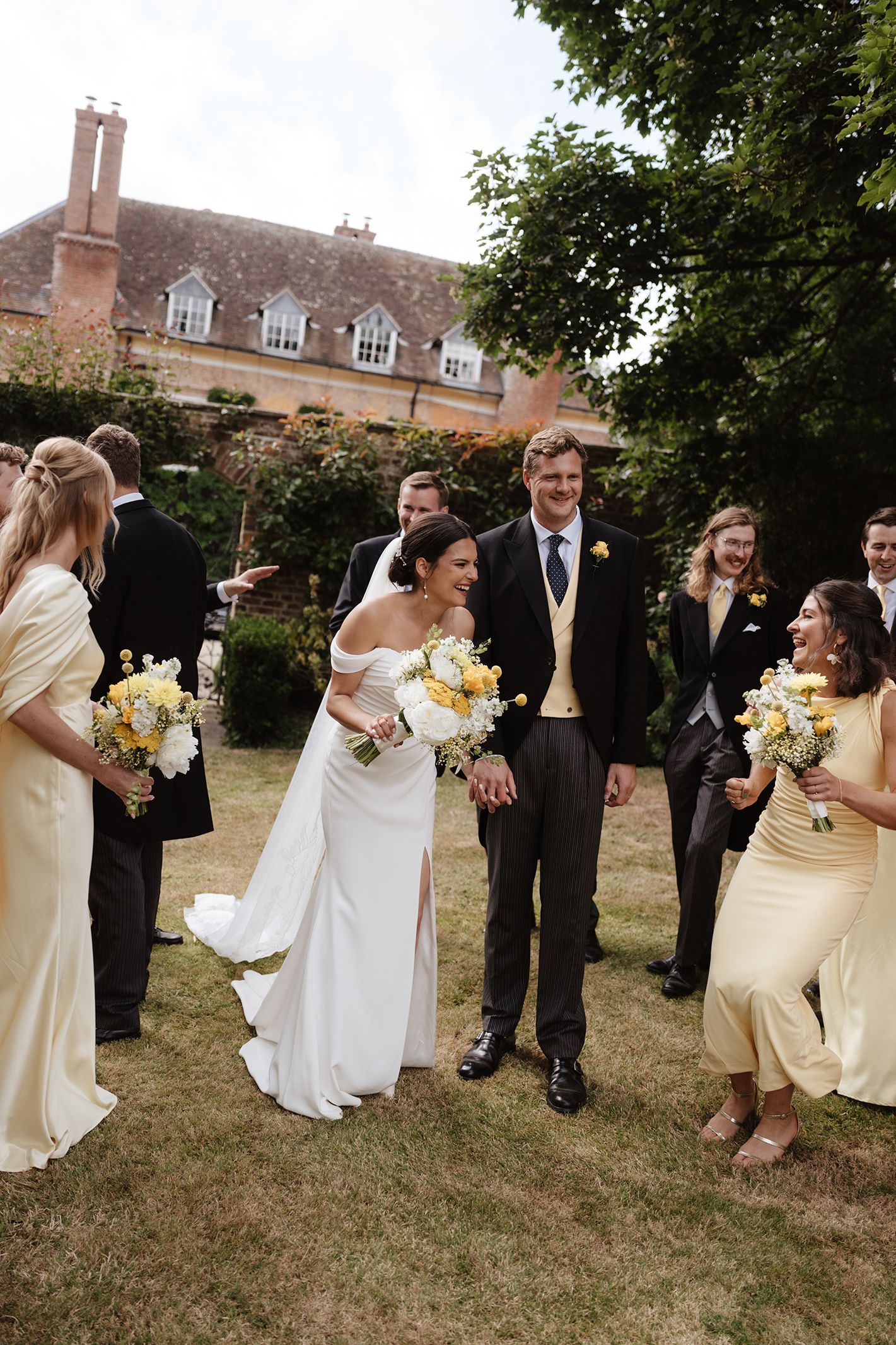Bride and groom walk through cheering wedding party on green lawn outside historic manor house.