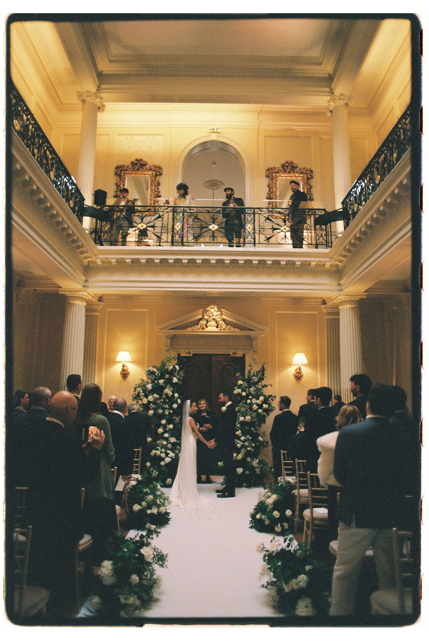 Elegant wedding ceremony in a grand ballroom with floral aisle decor, guests seated, couple at altar, musicians on balcony above.