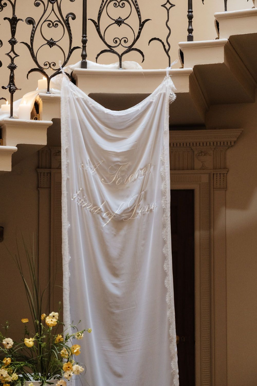 White wedding veil with embroidered text draped over ornate iron stair railing in elegant venue.