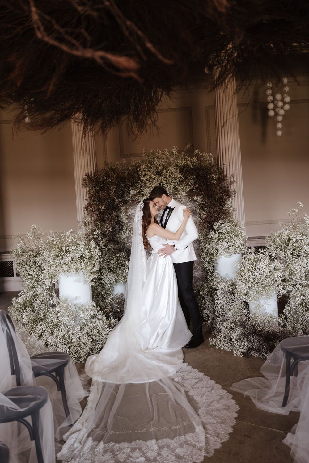 Bride and groom embrace at floral ceremony altar with baby's breath arrangements and candlelight in elegant ballroom.