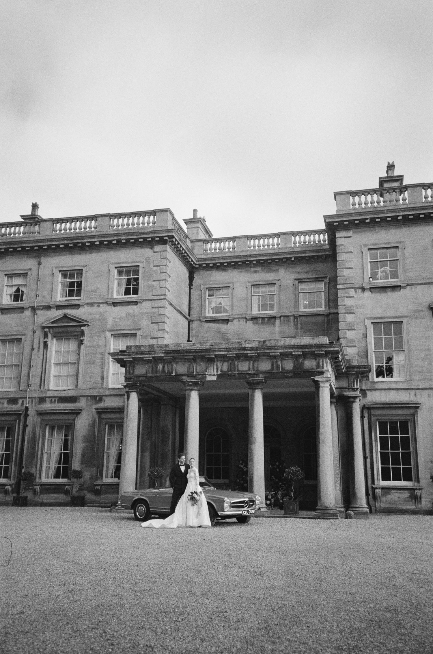 Black and white photo of a wedding couple posing in front of a grand historic mansion with classical columns and a vintage car.