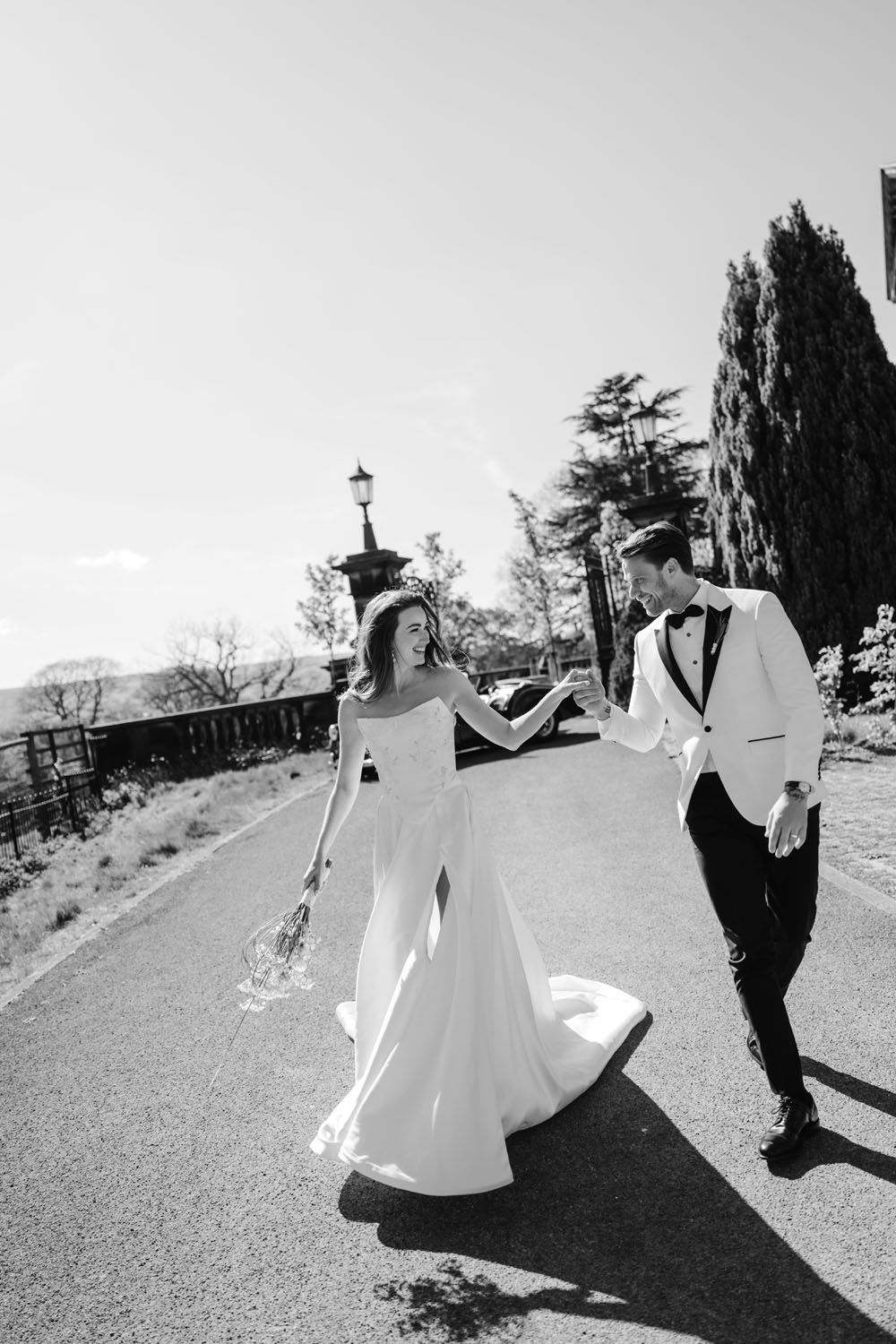 Black and white photo of a bride and groom holding hands, walking down a sunlit path on their wedding day.