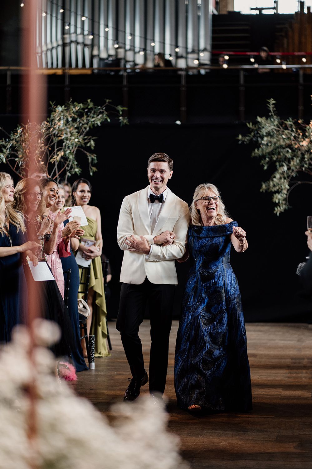 Groom walking down the aisle while holding his mams hand, she's in a blue dress and hes in a monochrome tux.