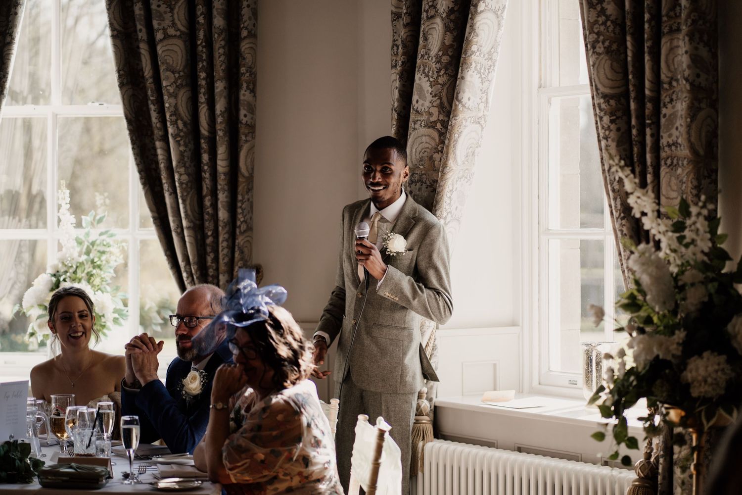 Best man introducing the start of wedding day speeches with windows behind him.
