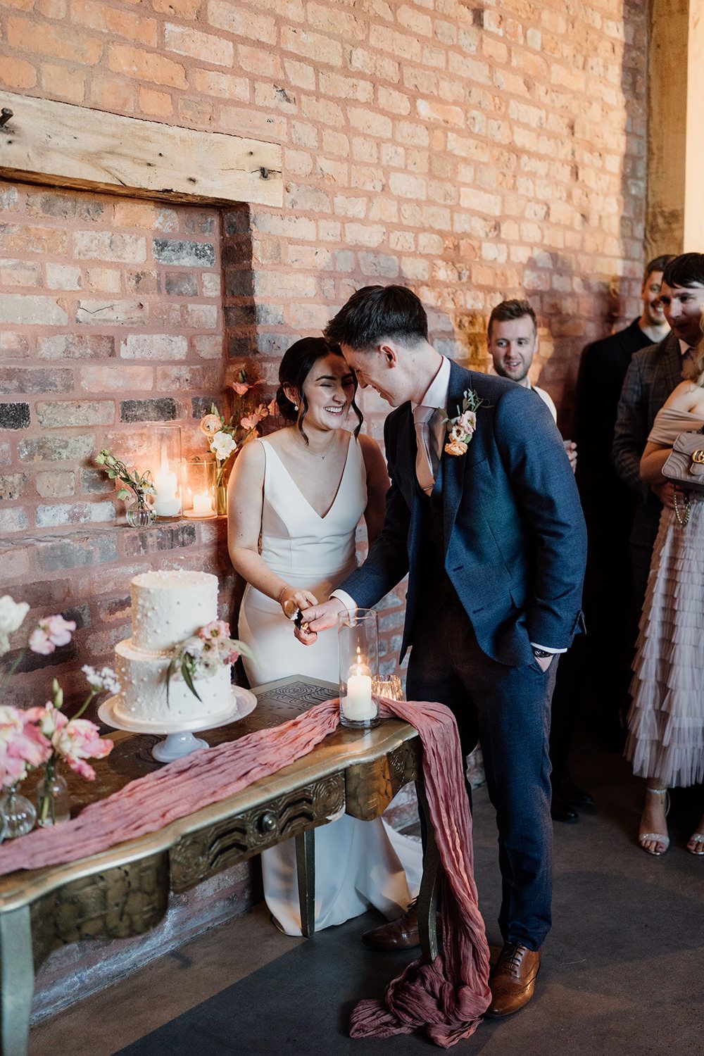 Bride and groom stood cutting their wedding cake with guests watching, the cake is on a table with flowers and candles.
