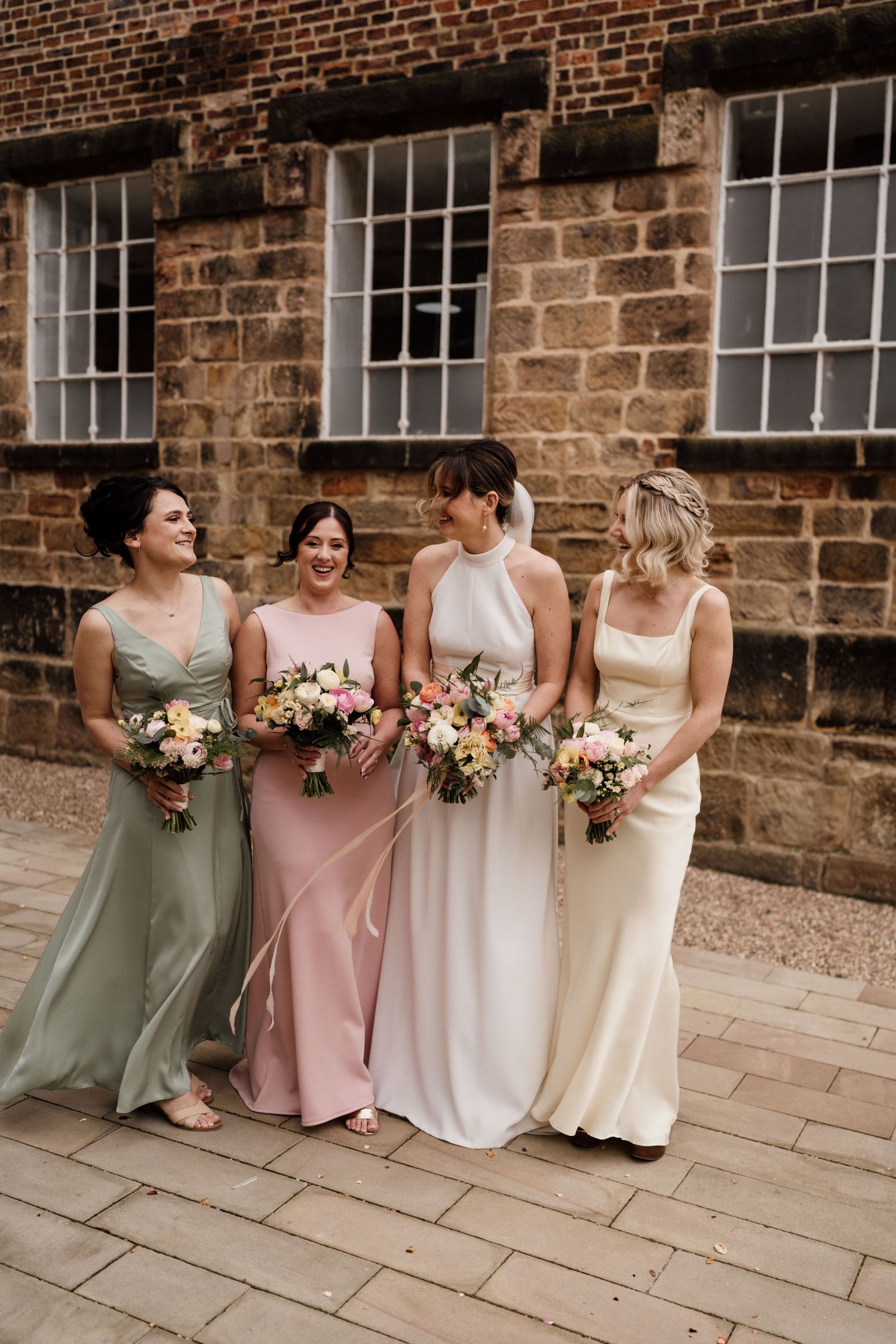 Four bridesmaids in pastel dresses hold pink floral bouquets against historic brick building.