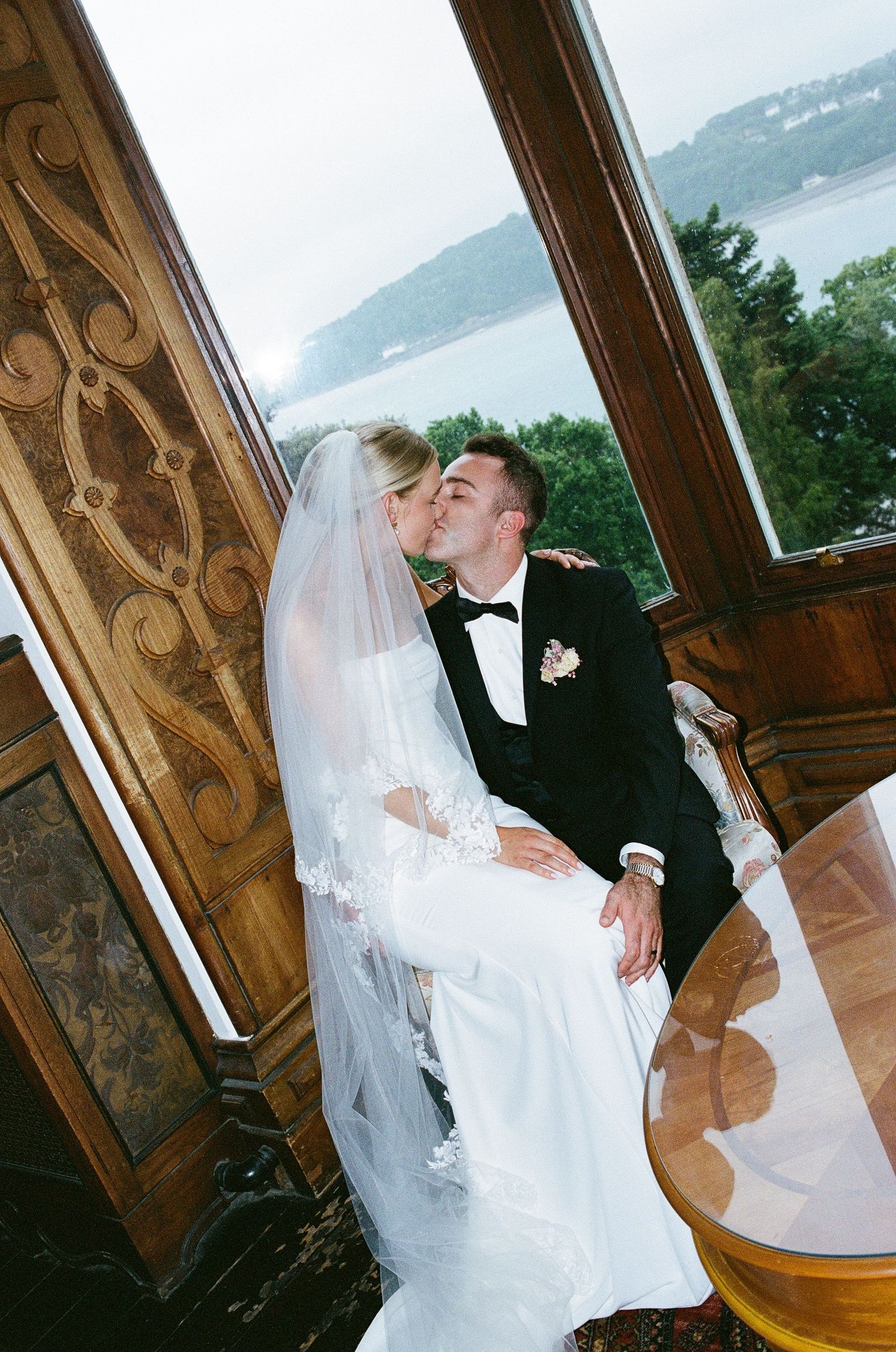 Bride and groom share a romantic kiss by a large window overlooking a scenic lake in a grand venue.
