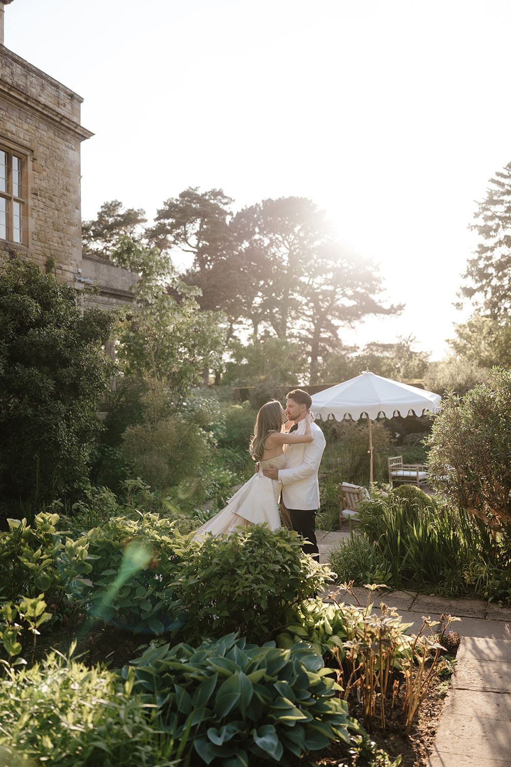 Romantic sunset wedding photo in a lush garden with sun flare and stone building in background.