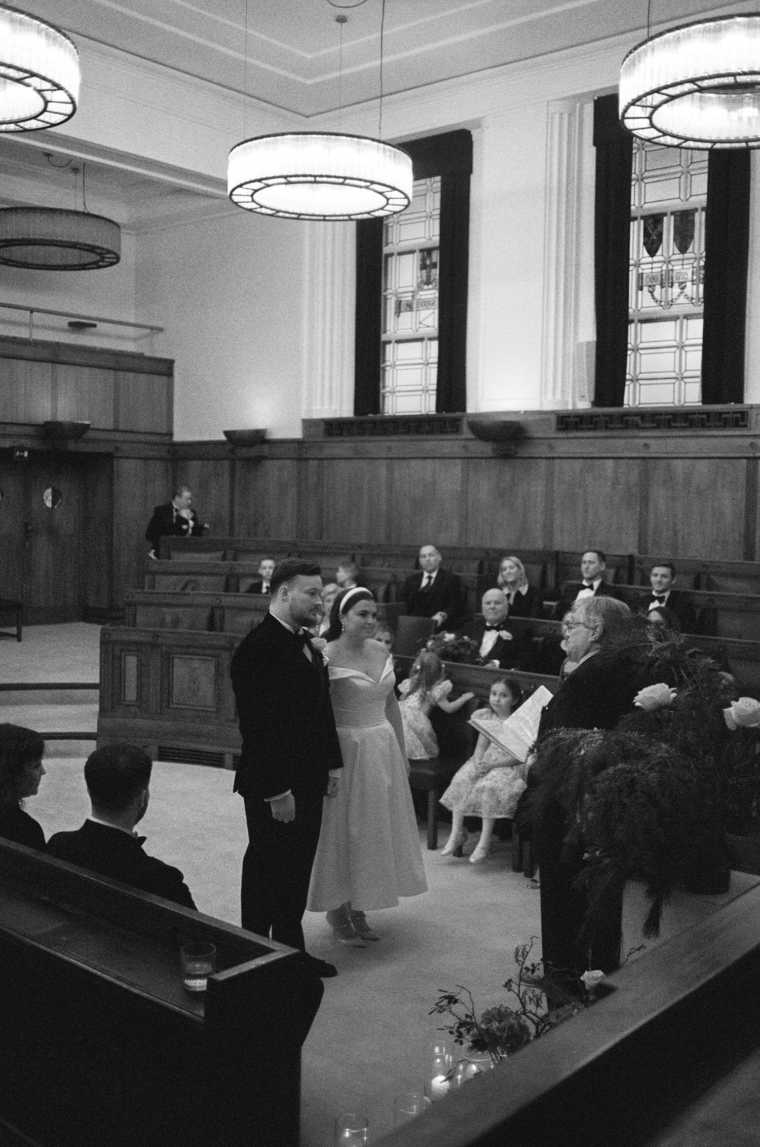 Black and white photograph of a wedding ceremony in a chapel with art deco light fixtures and wooden paneling.