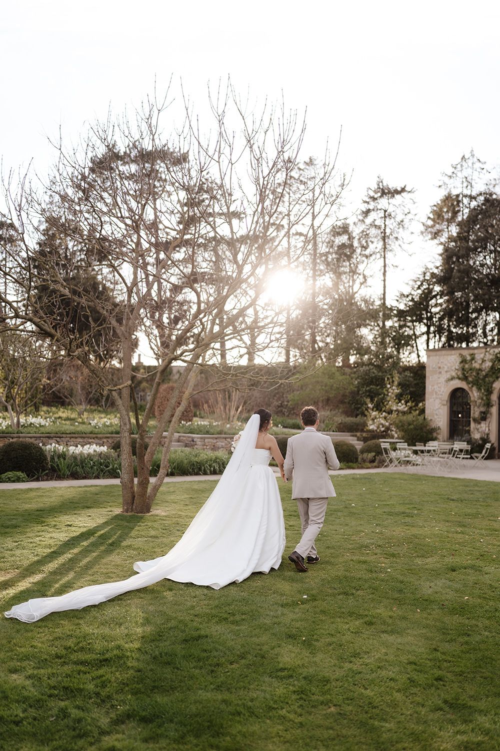 A couple walks hand in hand on a manicured green lawn at sunset with a long flowing white wedding dress trailing behind.