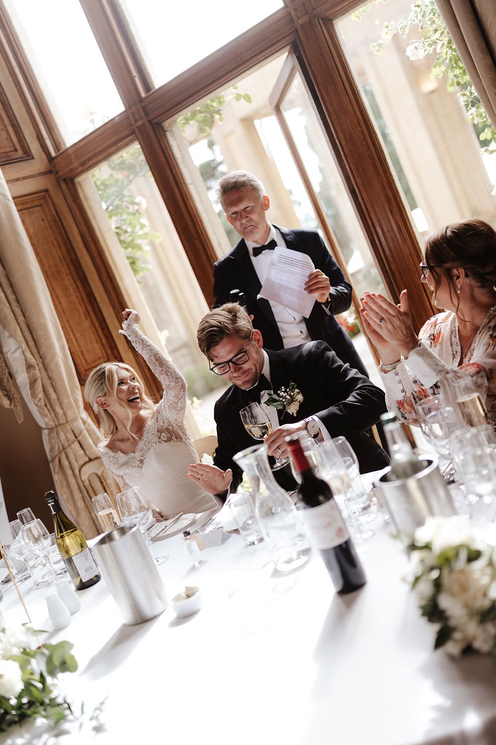Wedding guests laughing and enjoying themselves at an elegant reception with formal table settings and natural lighting.