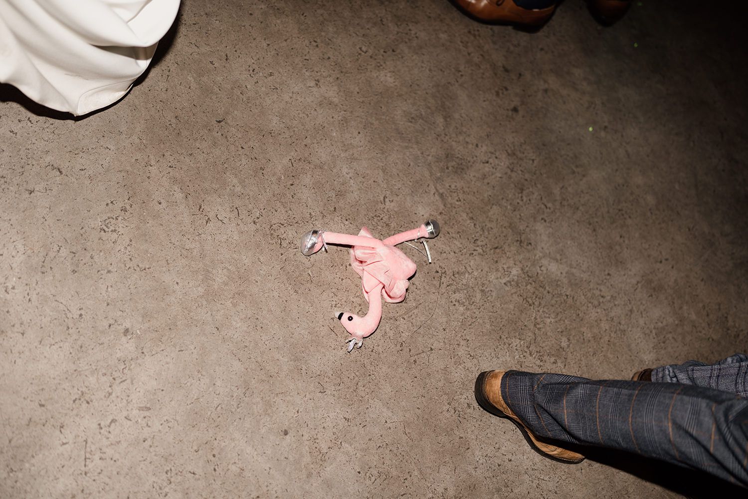 A toy pink flamingo on a grey concrete floor with wedding guests feet around.