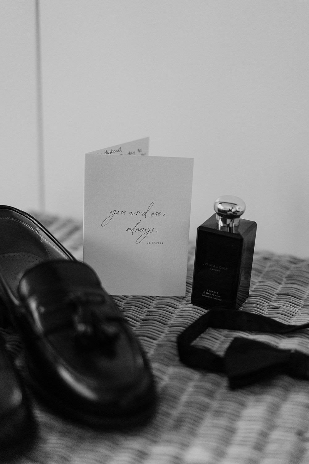 Black and white photo of dress shoes, perfume bottle and handwritten note on a textured fabric surface.