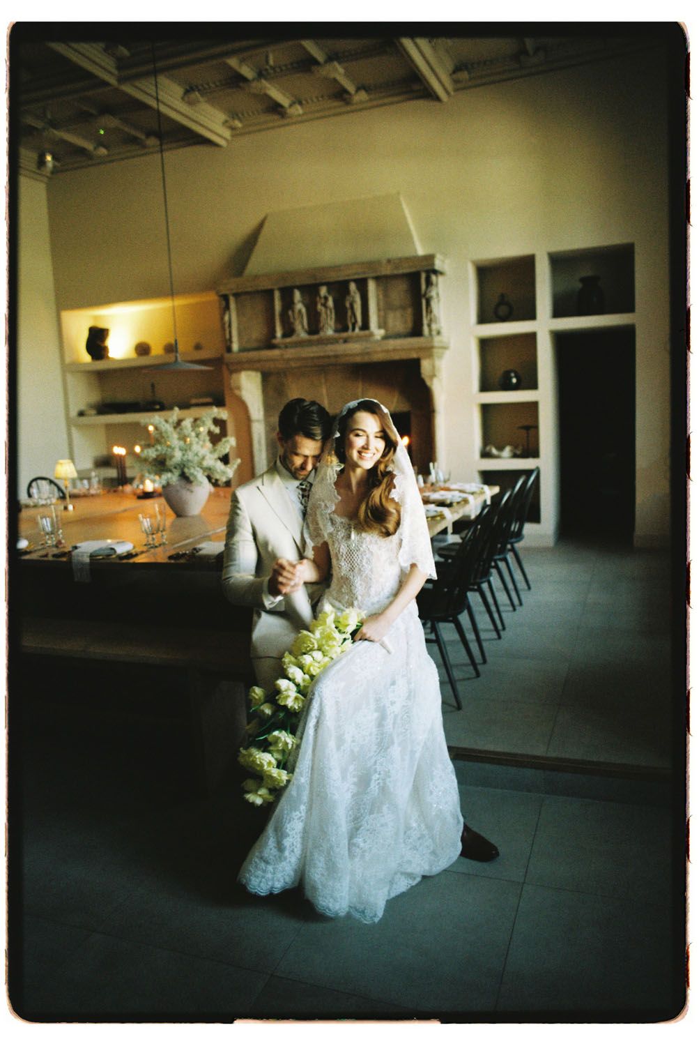Elegant bride in lace gown holding cascading green flowers poses with groom in rustic venue with stone fireplace.