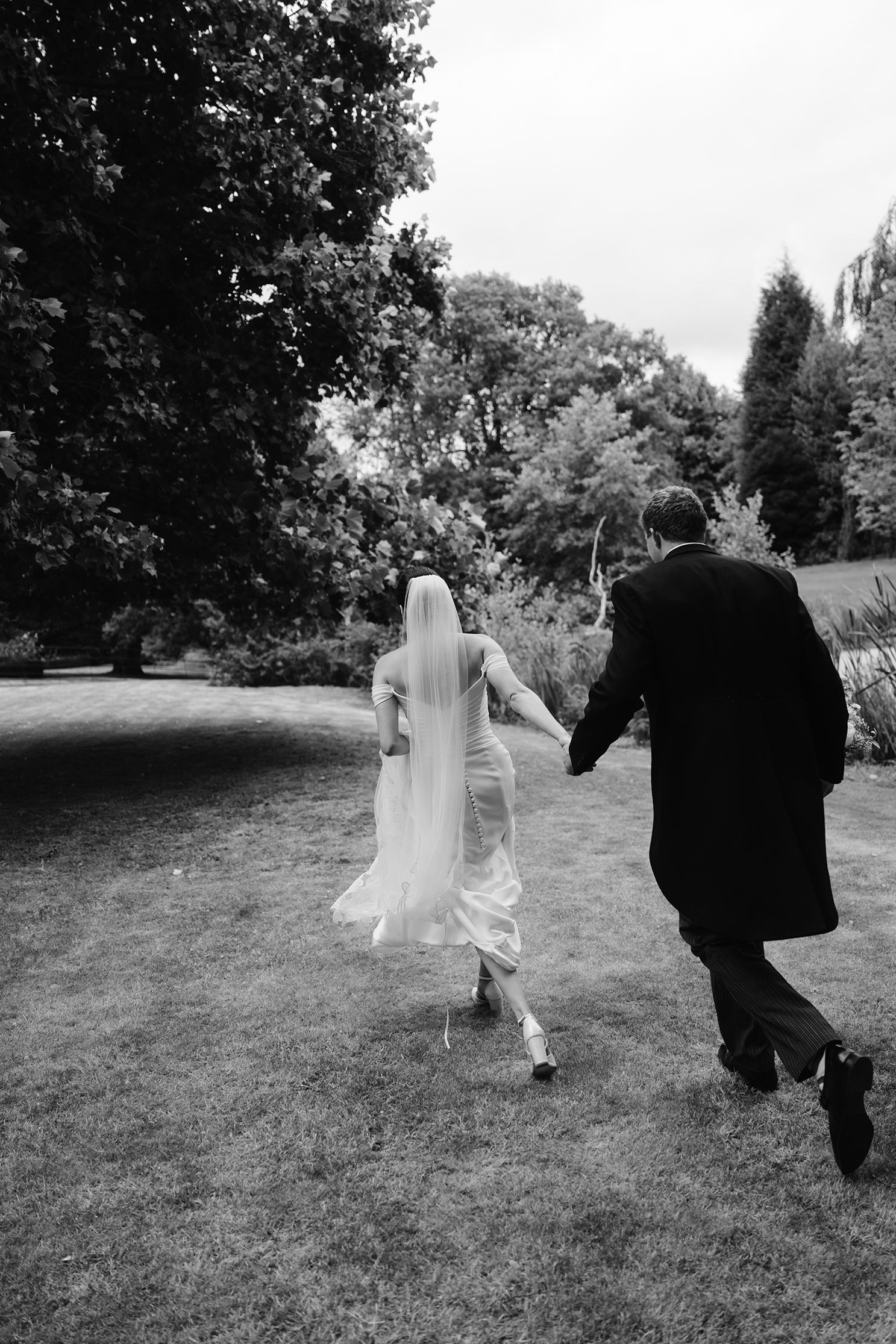 Bride and groom run hand in hand across lush garden lawn in classic black and white wedding photograph.