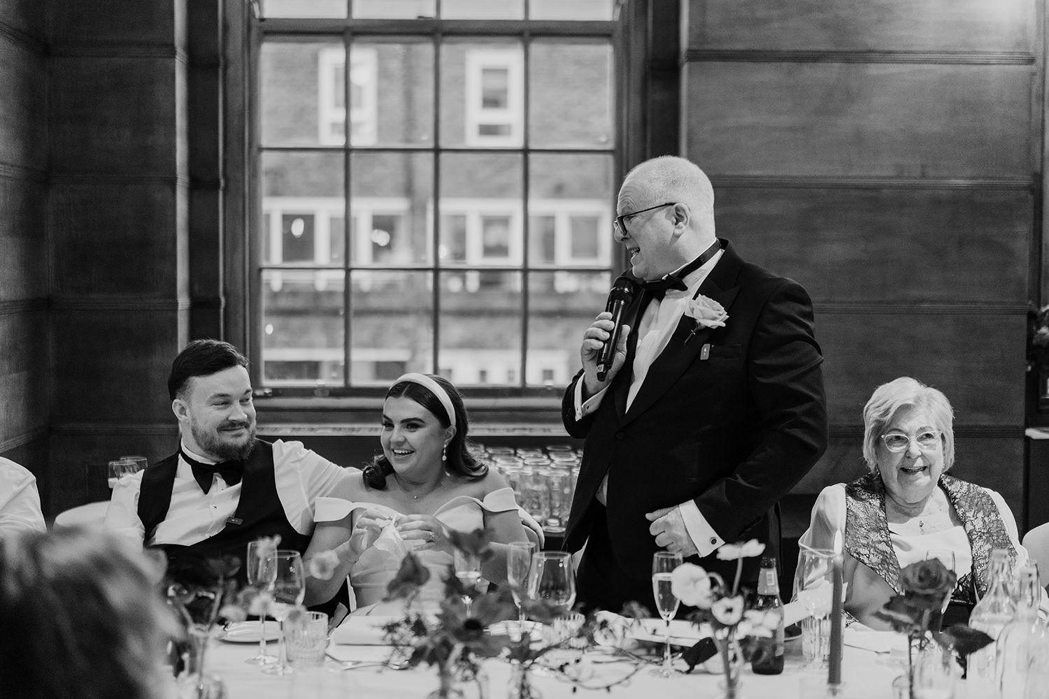 Black and white candid photograph of guests enjoying wedding reception dinner with speaker delivering a toast.