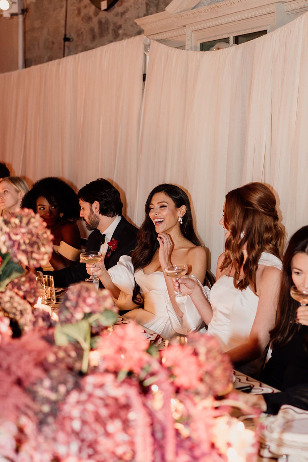 Wedding reception guests laugh and smile while seated at elegant tables decorated with pink floral centerpieces.