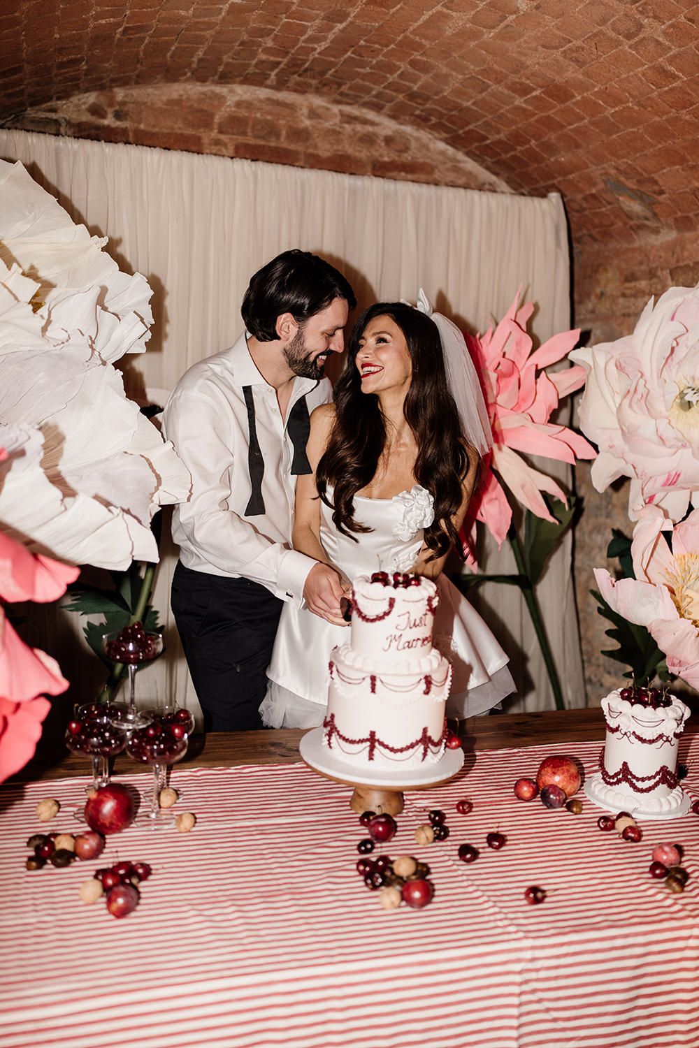 A couple shares a romantic moment by their wedding cake decorated with pink flowers and cherries on a striped tablecloth.