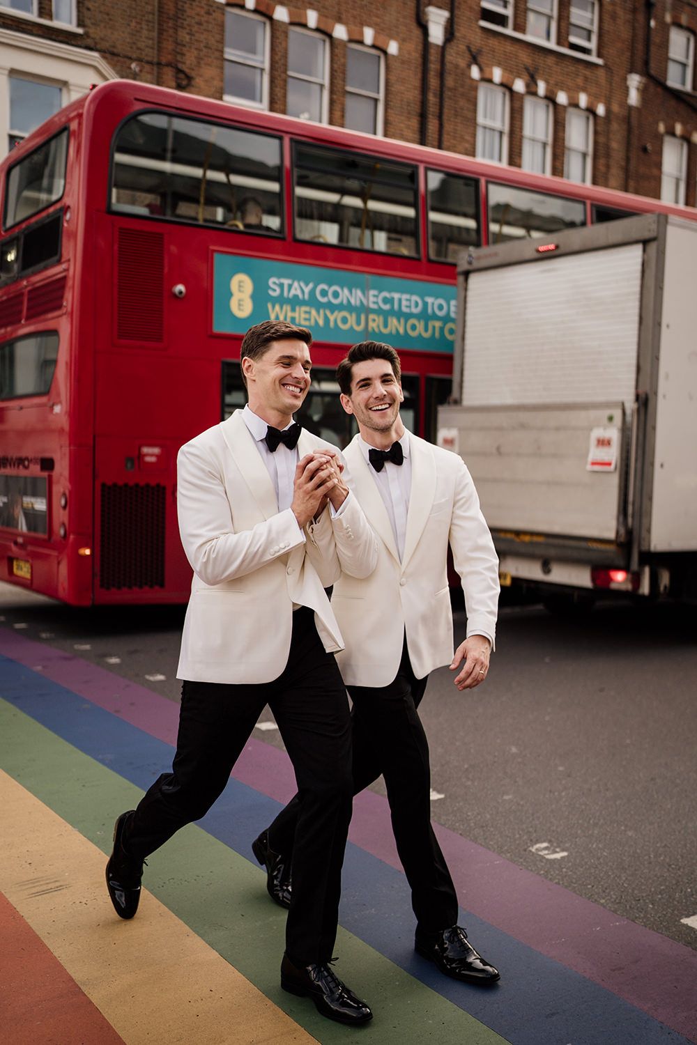 Two grooms wearing monochome suits as they cross a road with a red london bus behind them and a colourful floor.