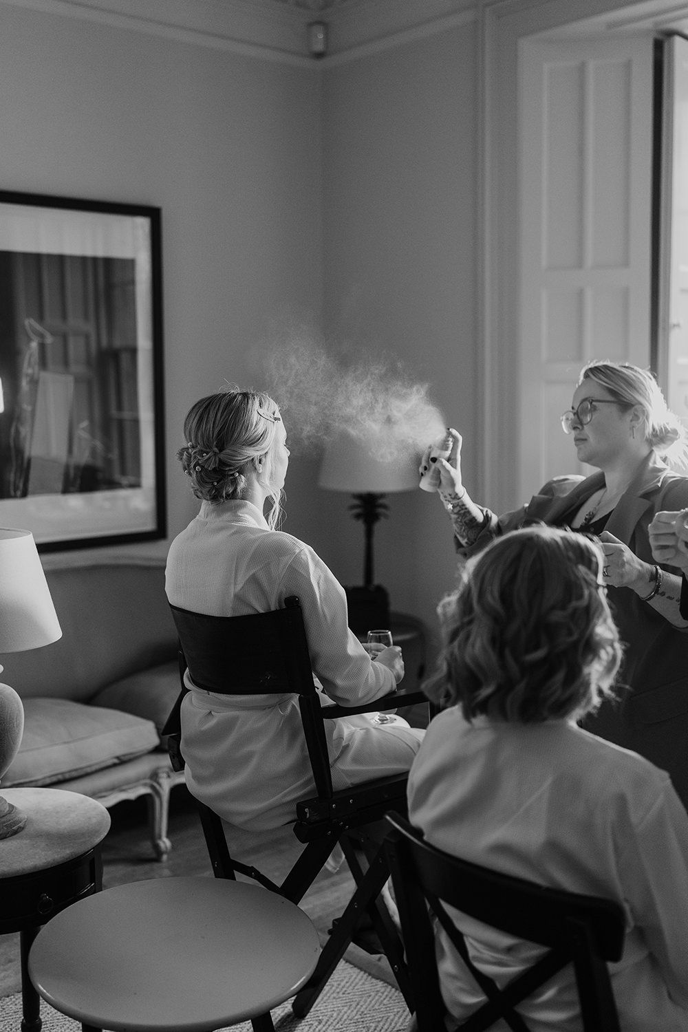 Makeup artist applying a spray to brides finished makeup while she is sat on a chair.