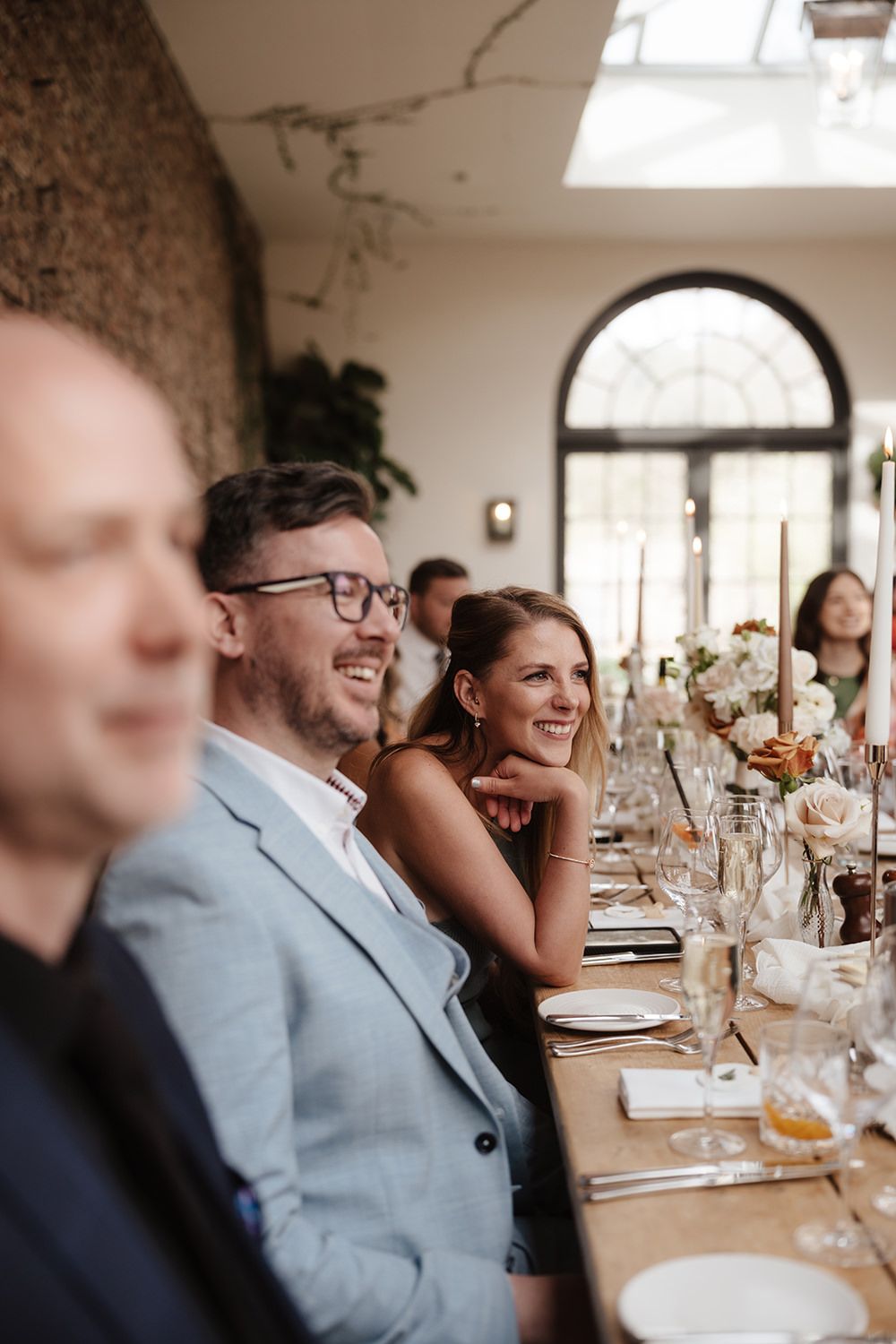 Wedding guests share joyful laughter at an elegant reception dinner with white floral centerpieces and formal place settings.