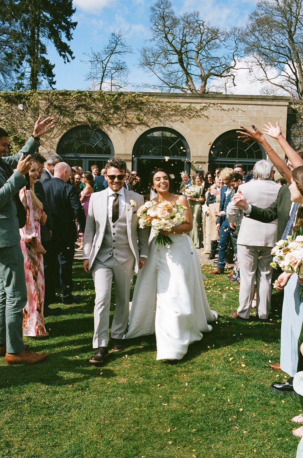 Wedding guests shower flower petals on a newlywed couple walking along a garden path with arched architecture in background.