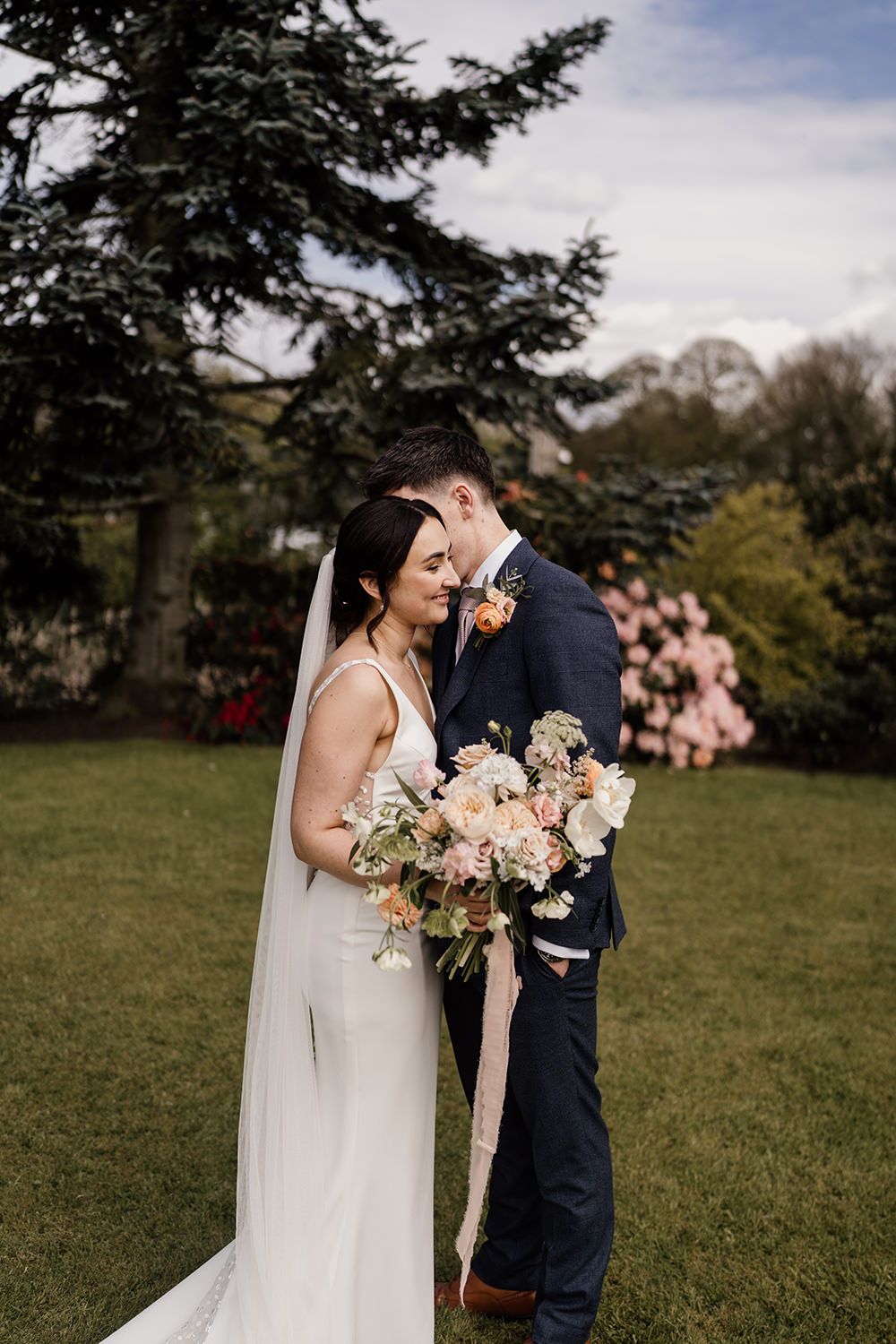 Groom and Bride stood while groom kisses bride on cheek, with gardens at The Holford Estate in the background.