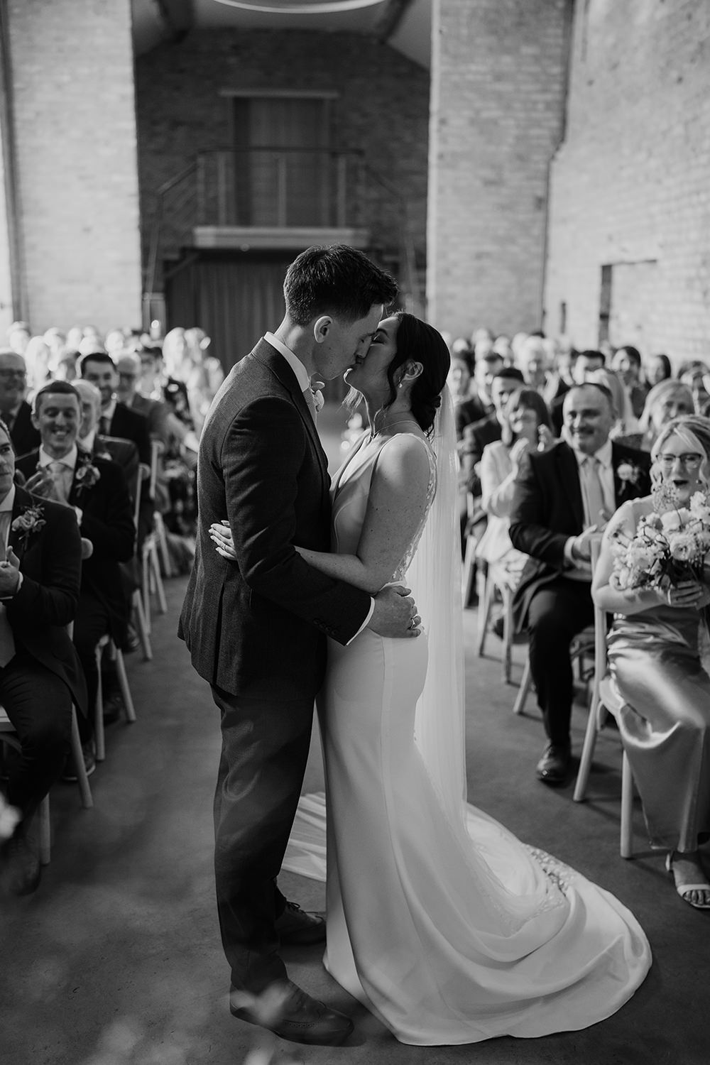 Bride and groom stood kissing at wedding ceremony with guests in the background.
