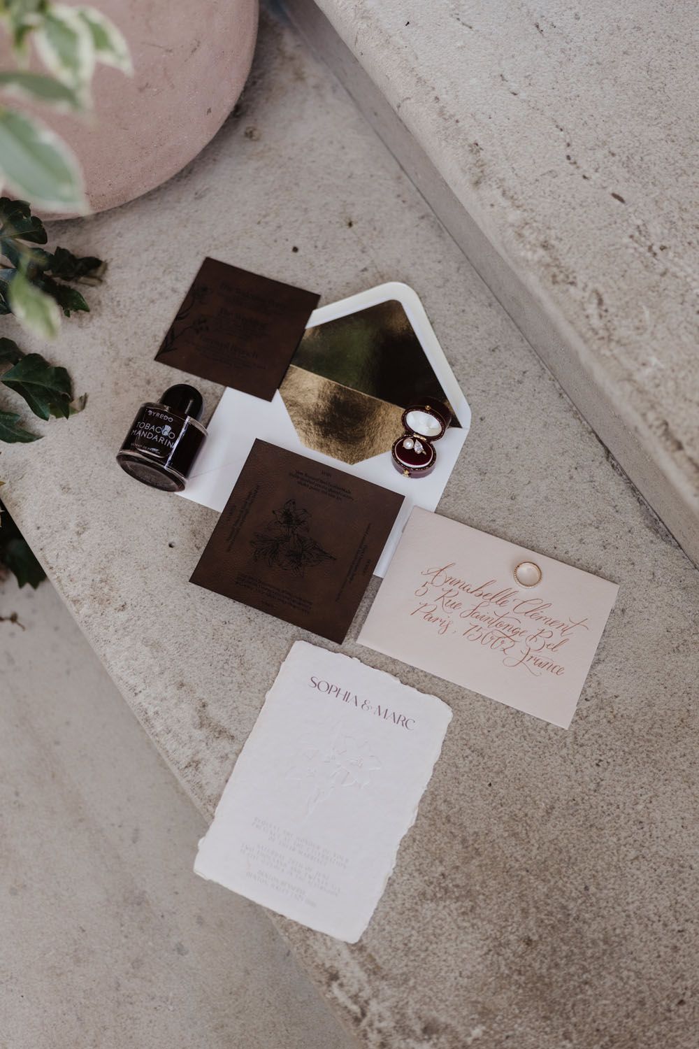 Elegant wedding stationery flat lay with dark brown velvet invite, gold-lined envelope, ring box, and ink bottle on concrete.