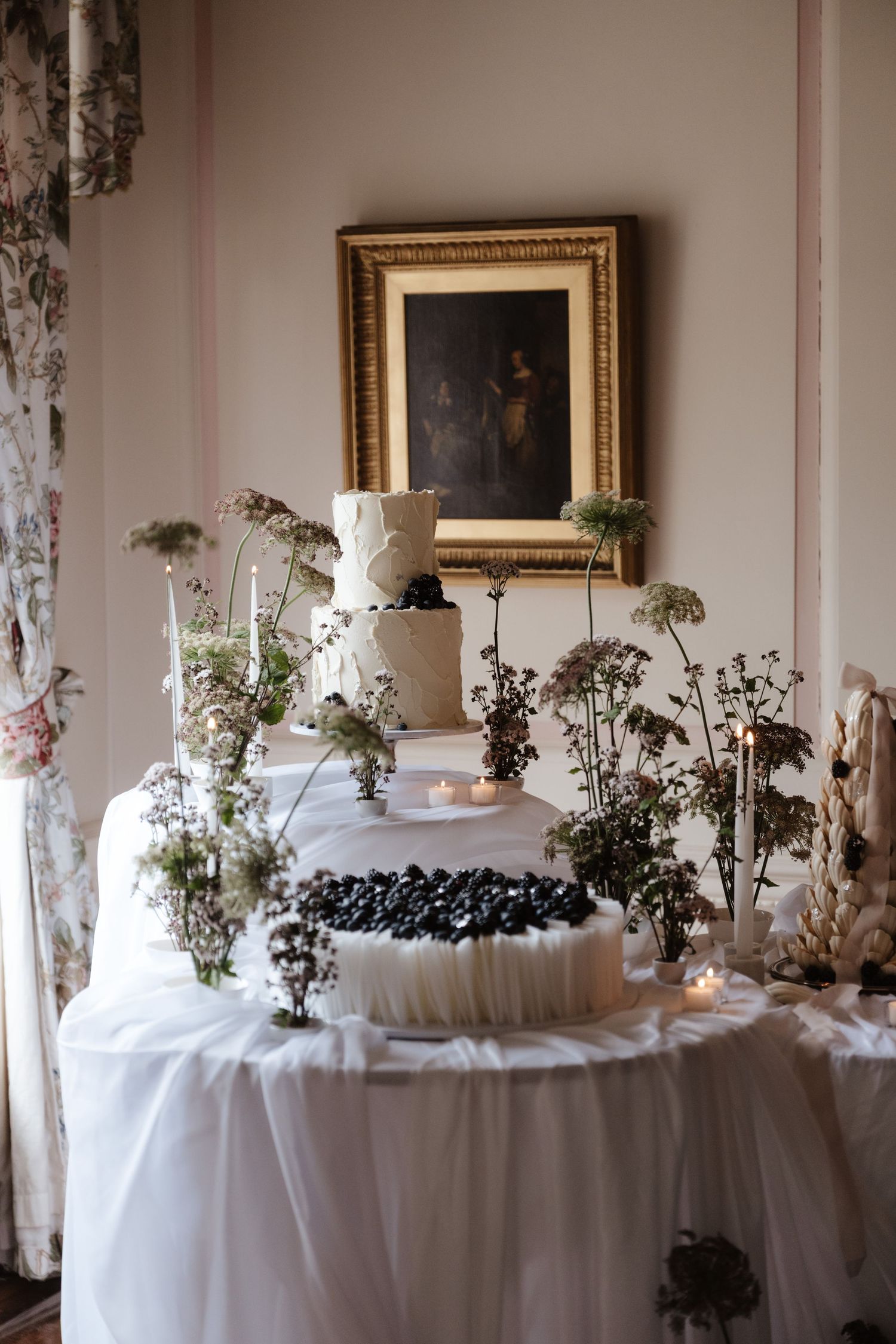 White tiered wedding cake with blueberries surrounded by wildflowers and candles beneath gold-framed portrait.