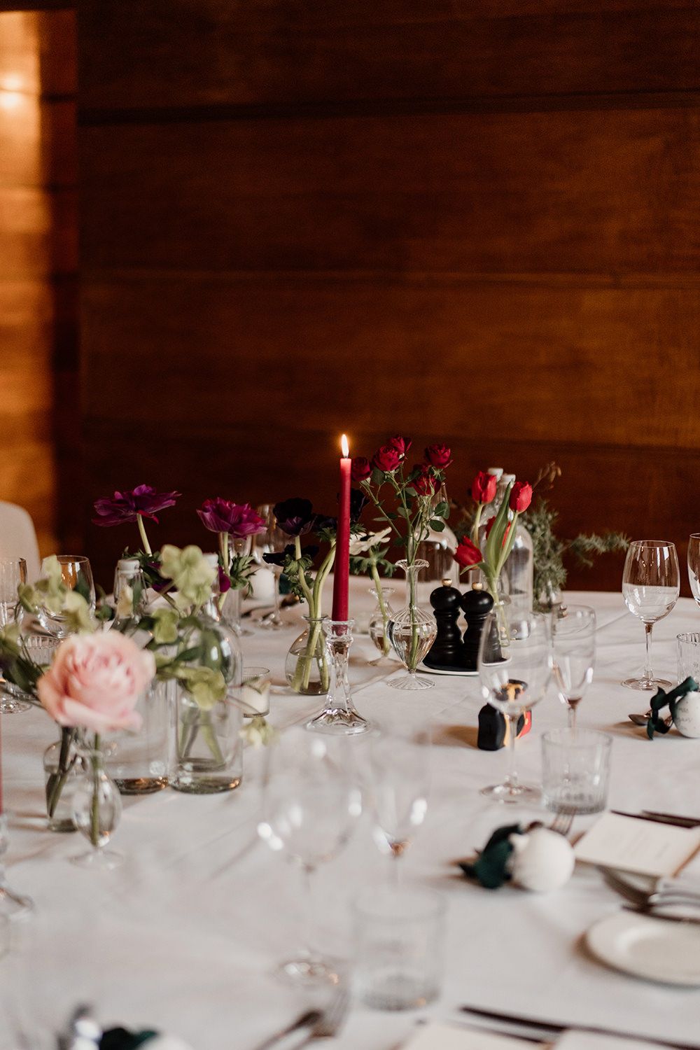 Elegant table setting with pink and purple flowers, red candles, and wine glasses on white tablecloth against wooden wall.