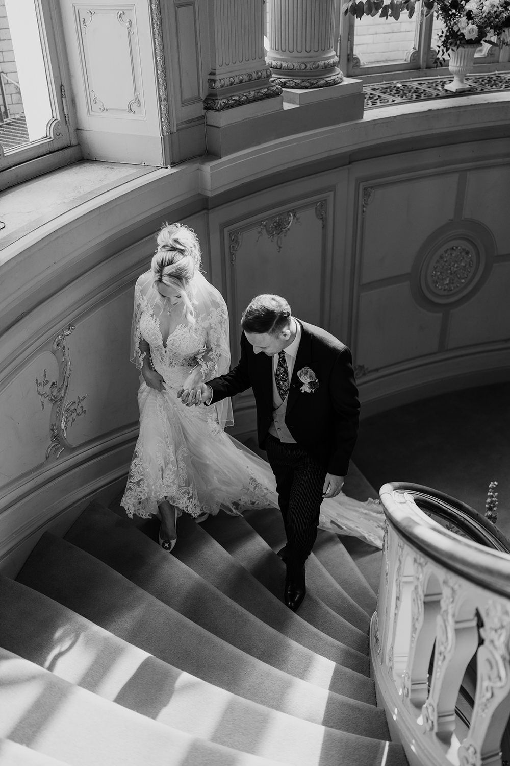 A black and white wedding photo of a couple descending an elegant curved staircase with ornate architectural details.