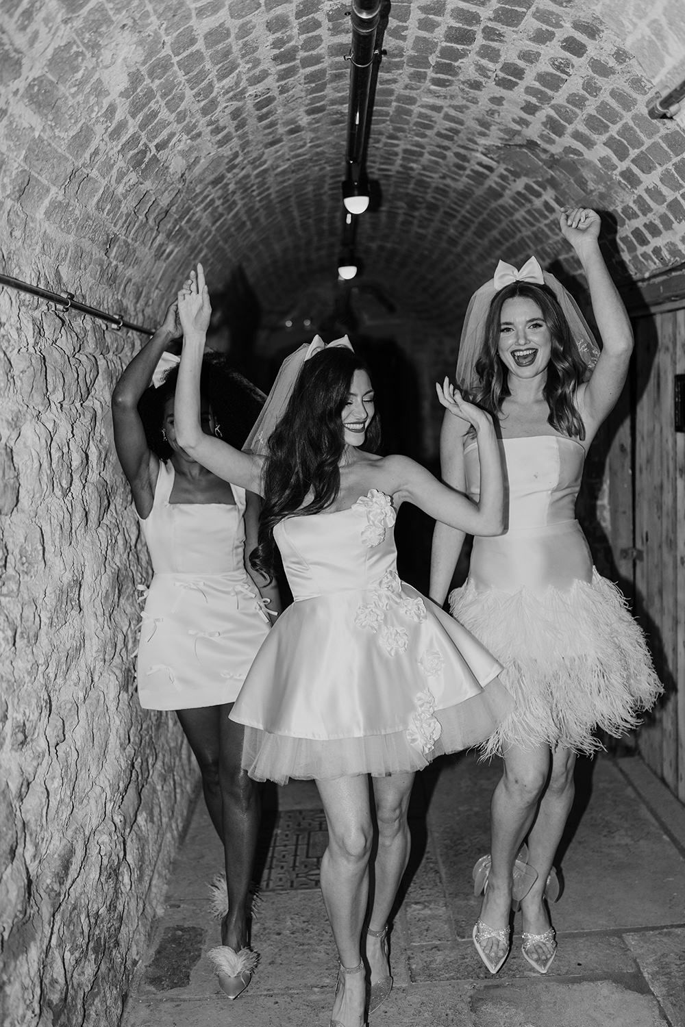 Three figures in white dresses dancing joyfully inside a brick tunnel with dramatic lighting.