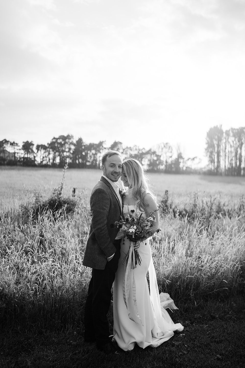 Bride and groom touching heads and smiling while the bride holds her flowers, stood in a field with a sunset behind.