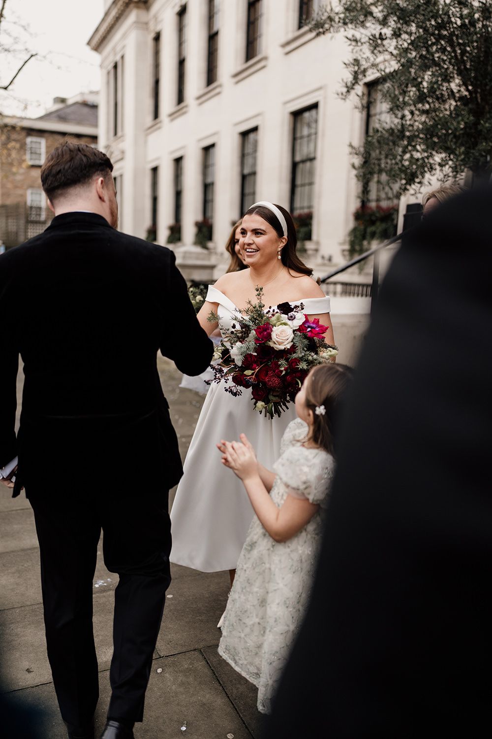 Beautiful wedding scene outdoors with bride holding burgundy bouquet standing on stone steps wearing off-shoulder gown.