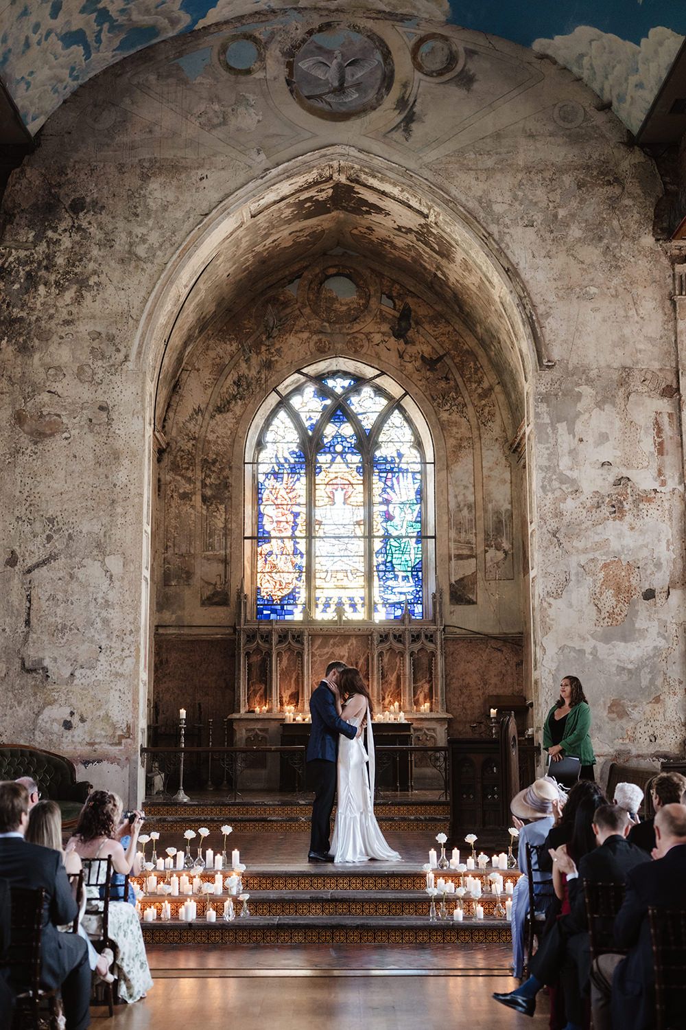 A wedding ceremony takes place in a historic chapel with stained glass windows and candlelight illuminating the stone interior.
