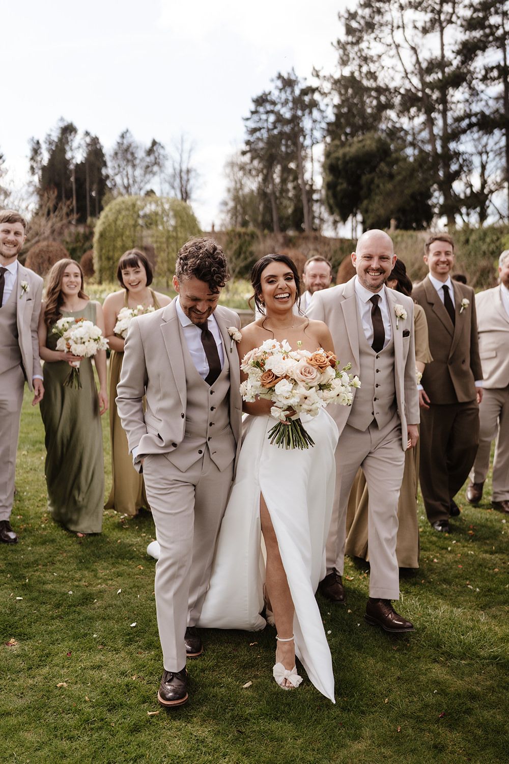 Wedding party wearing beige suits and olive dresses walk together on grass while celebrating joyfully outdoors.