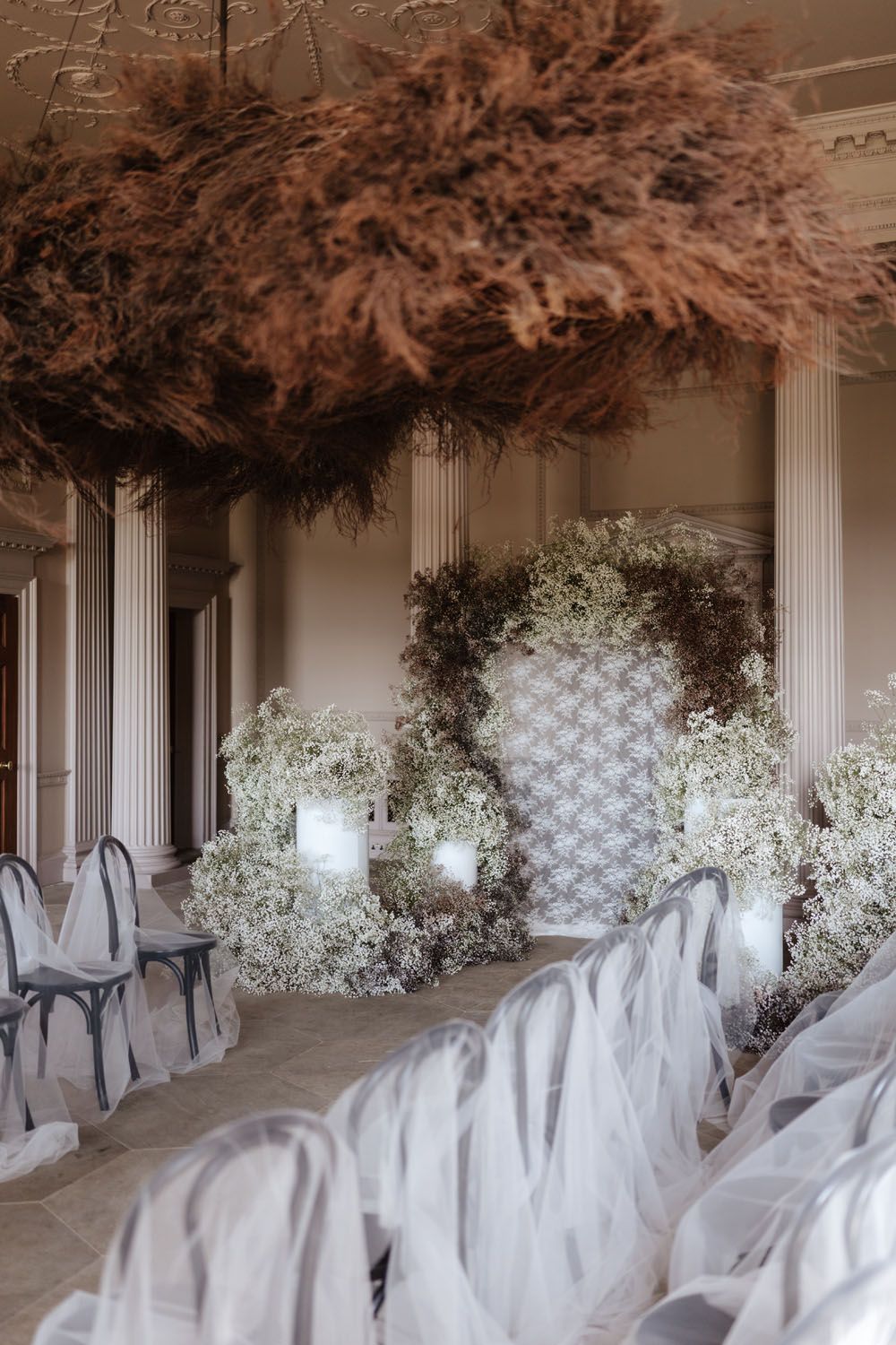 Elegant wedding ceremony setup with white chairs, floral arch, baby's breath, and dramatic dried pampas grass ceiling installation.