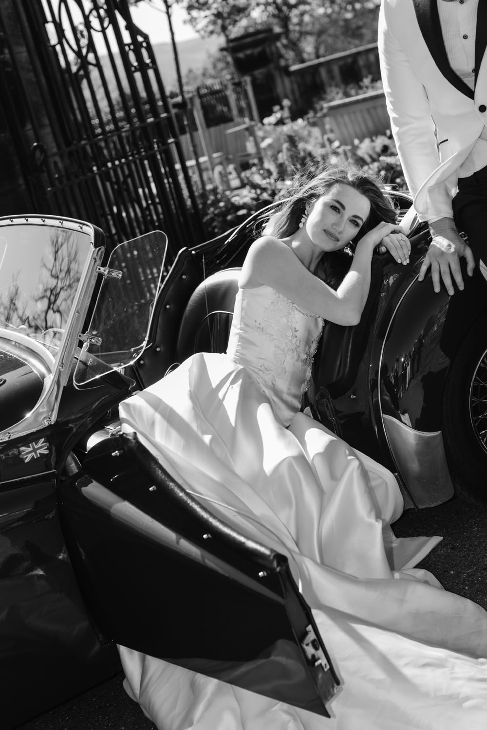 Black and white photo of a bride in a white gown sitting elegantly in a vintage sidecar motorcycle beside her groom.