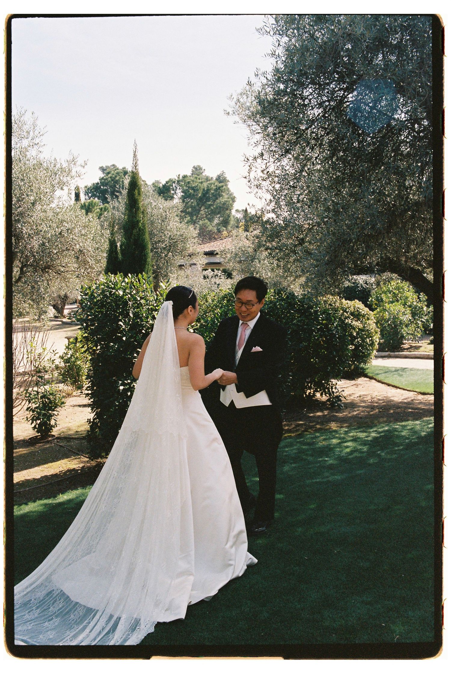 Bride and groom share a tender first look moment in a lush garden, her long veil trailing gracefully on the grass.