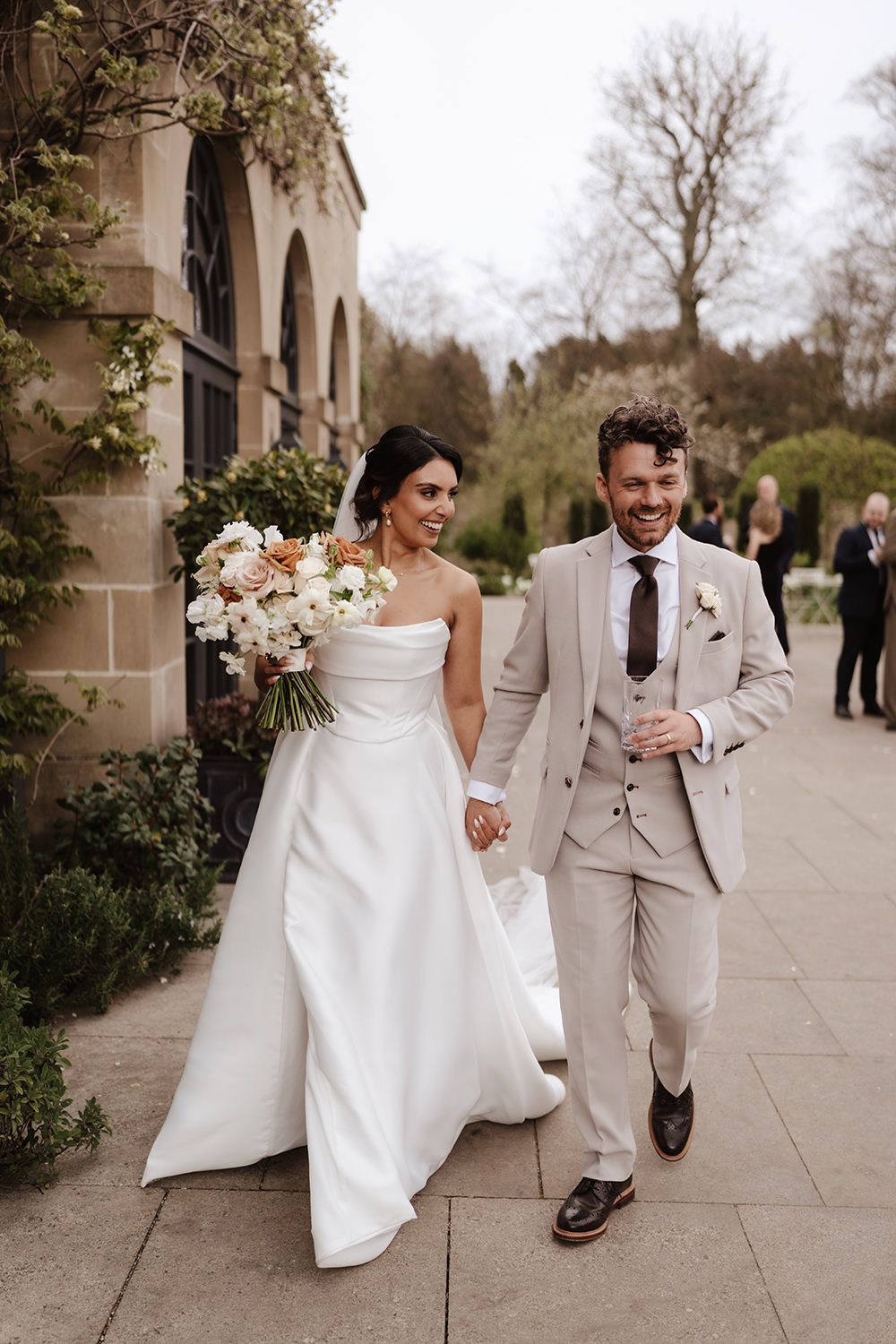 A wedding couple walks together on a stone path, with the bride in white and groom in beige holding hands.