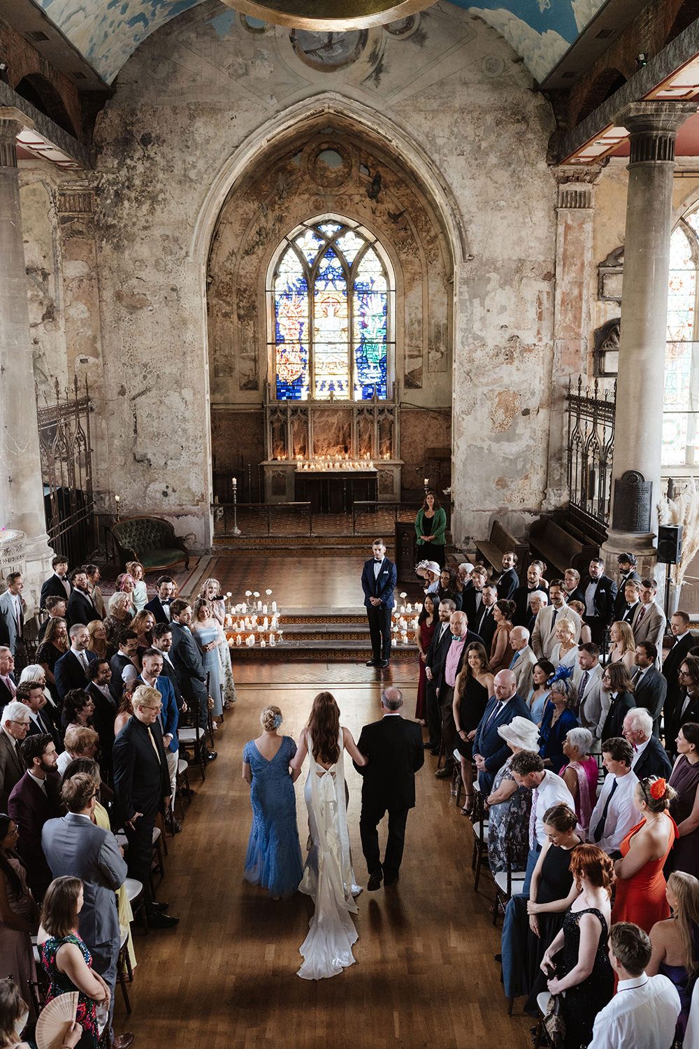 Wedding guests gather in a rustic church hall with stained glass windows and weathered stone walls.