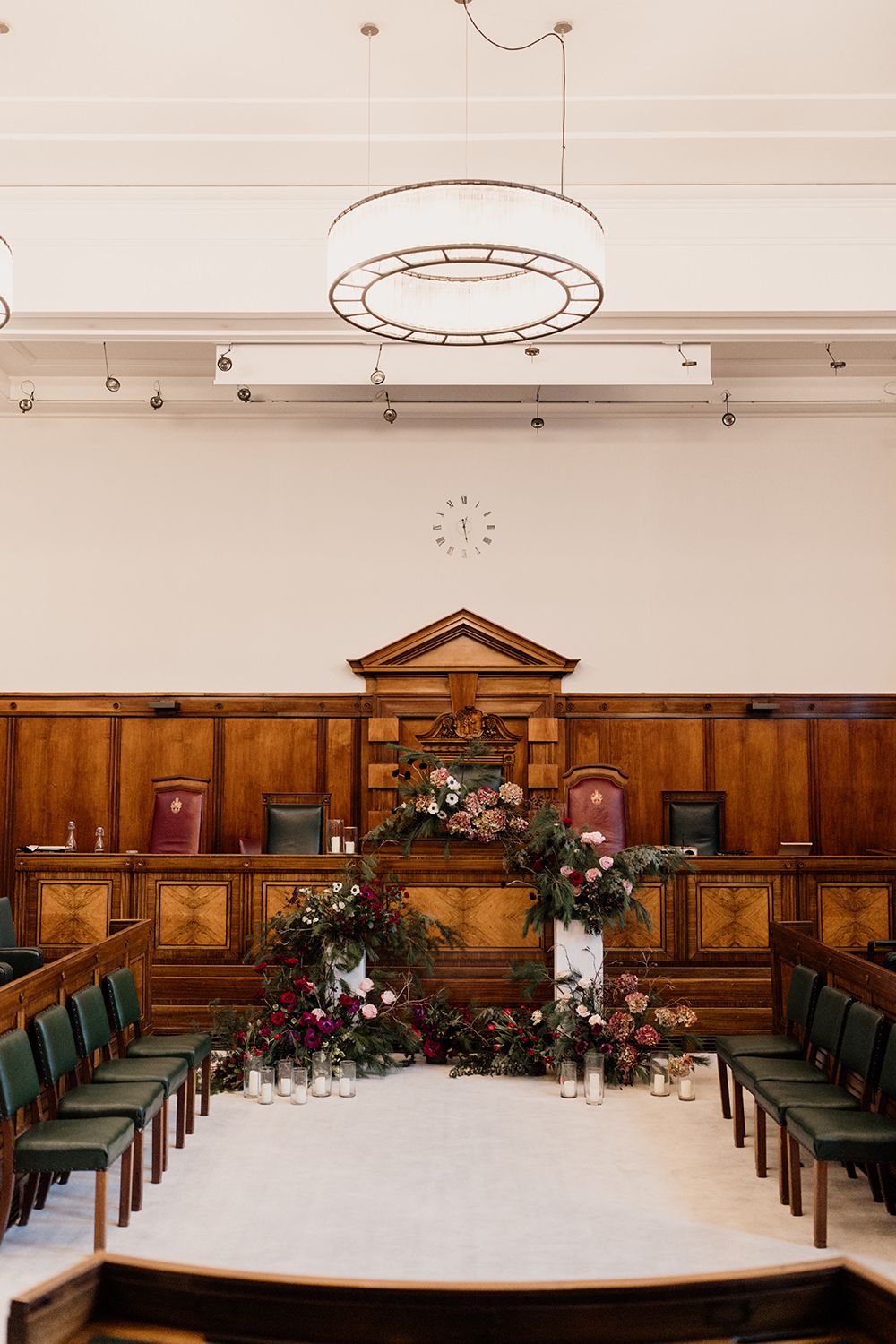 Elegant ceremony room with wooden paneling, green chairs, and ornate floral arrangements on a raised platform.