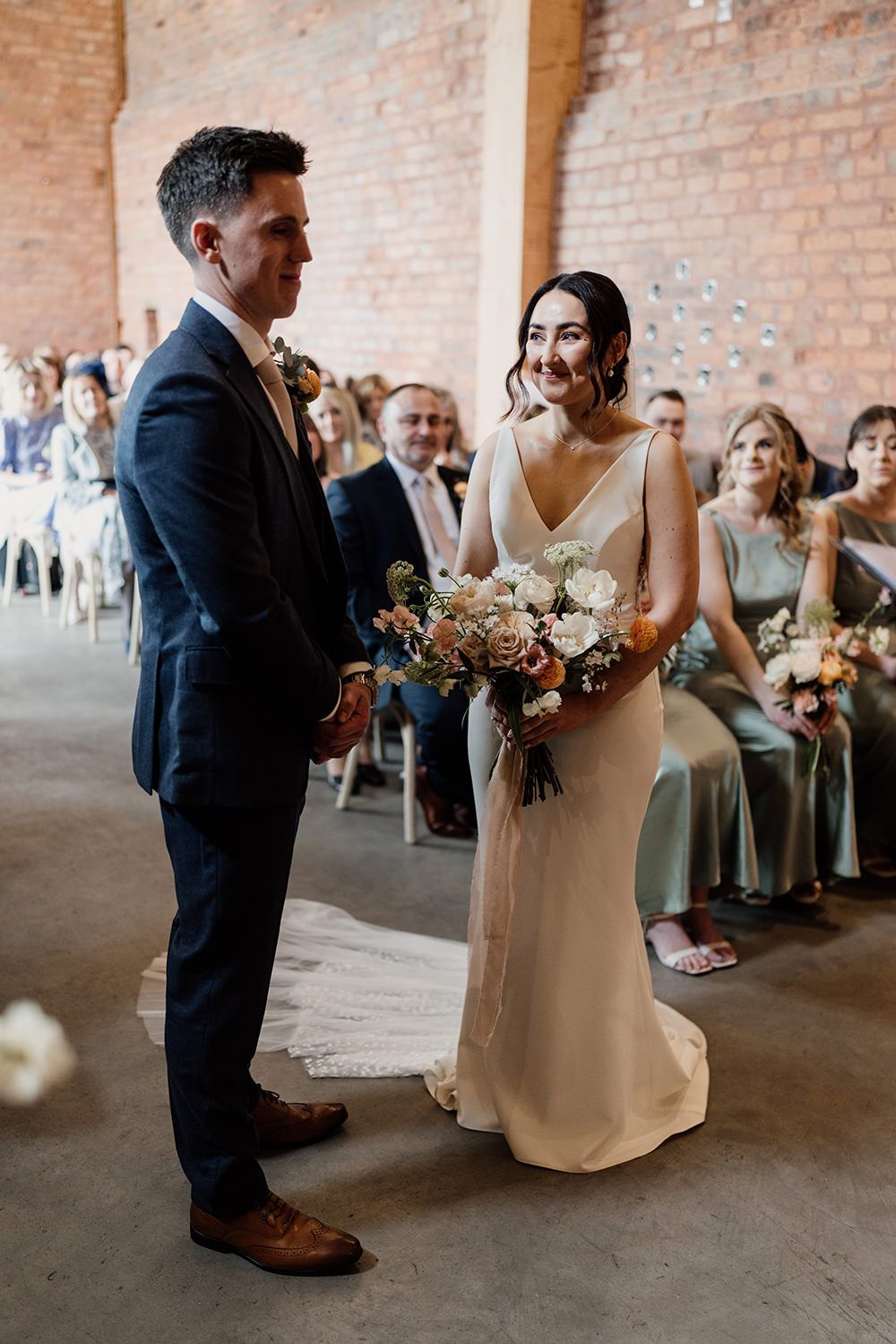 Bride stood smiling at her groom while holding flowers during wedding ceremony at The Holford Estate.
