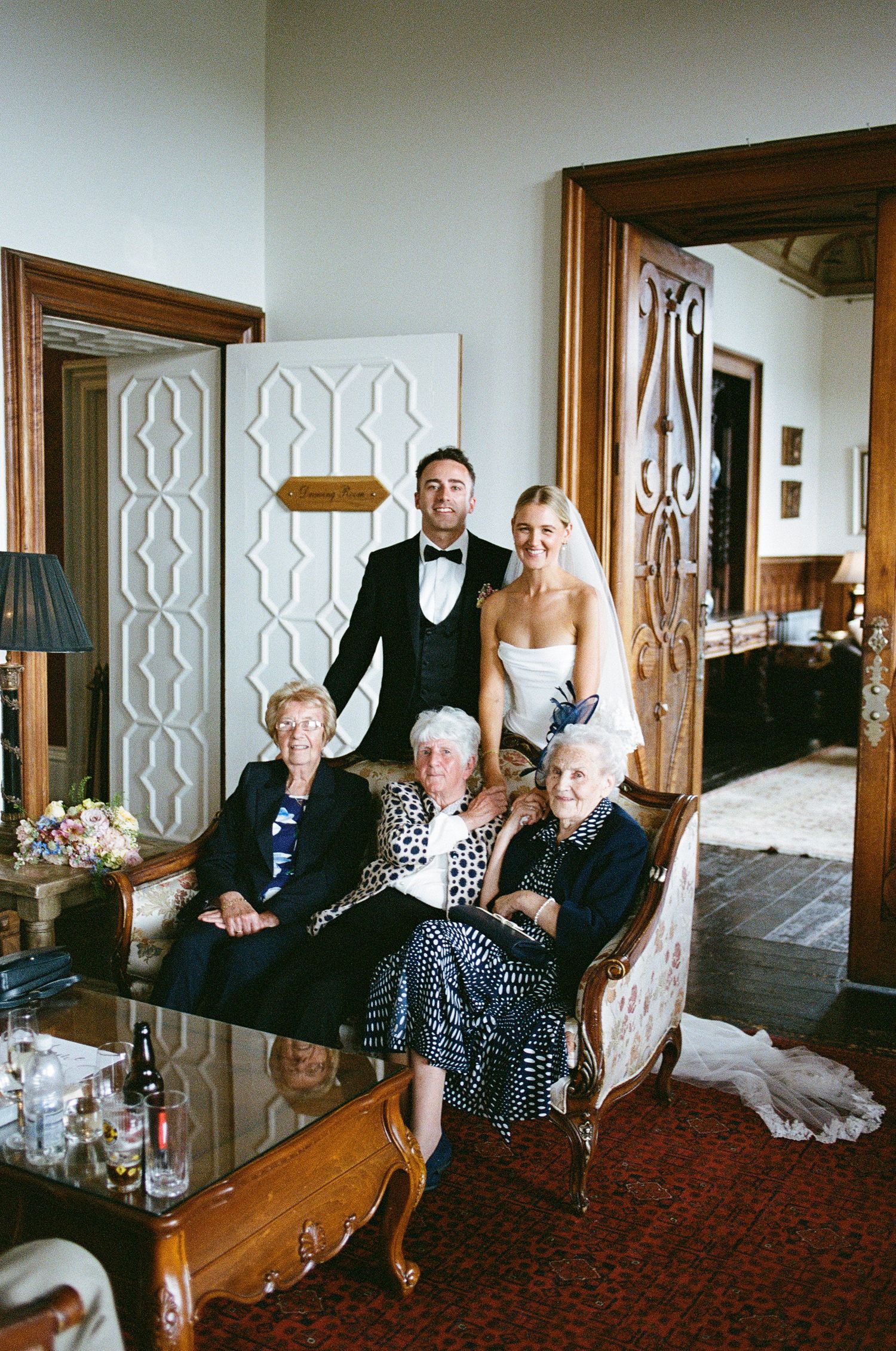 Bride and groom pose with three elderly seated women in an elegant room during a wedding celebration.