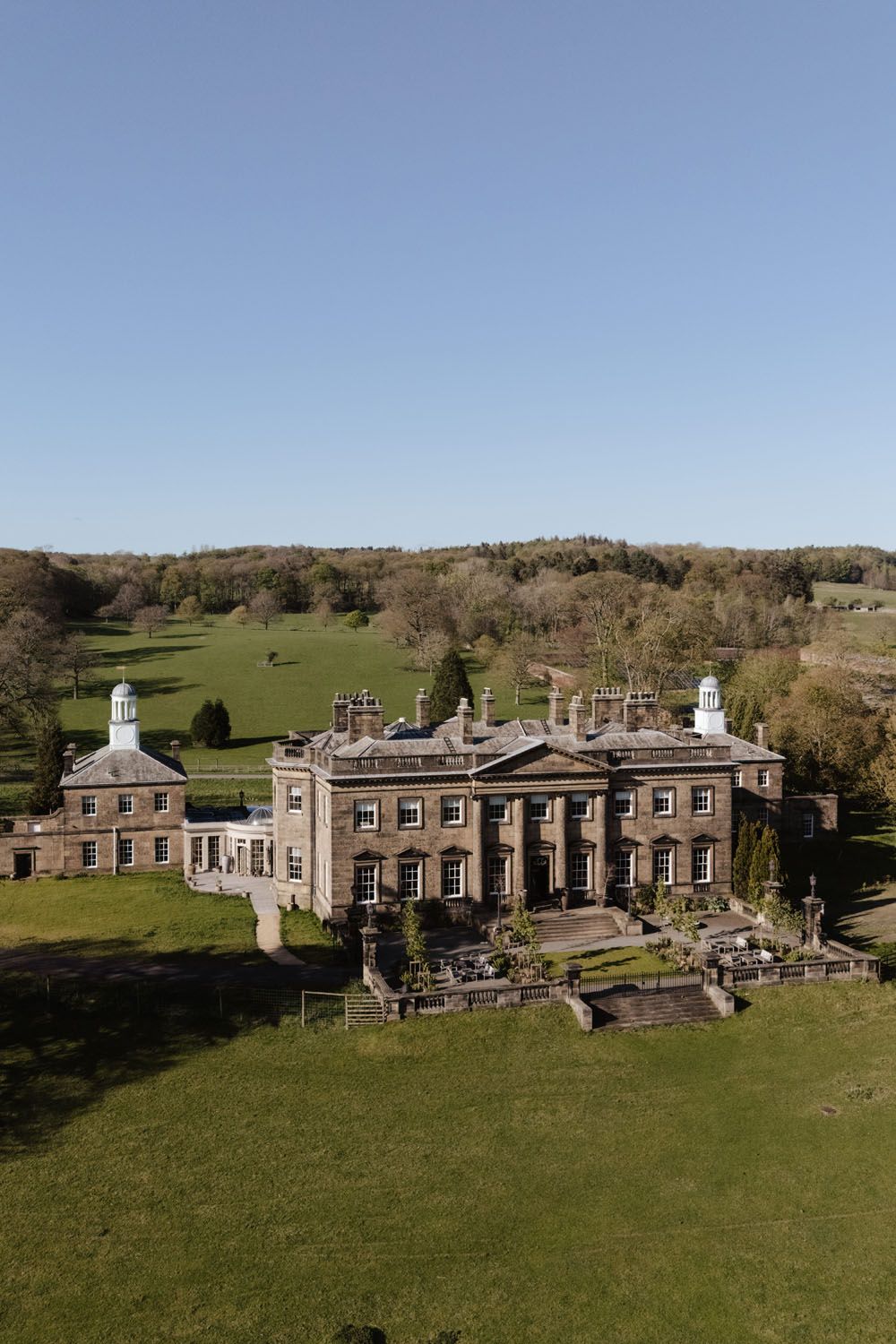 Aerial view of a grand Georgian country house surrounded by lush green lawns and woodland under a clear blue sky.
